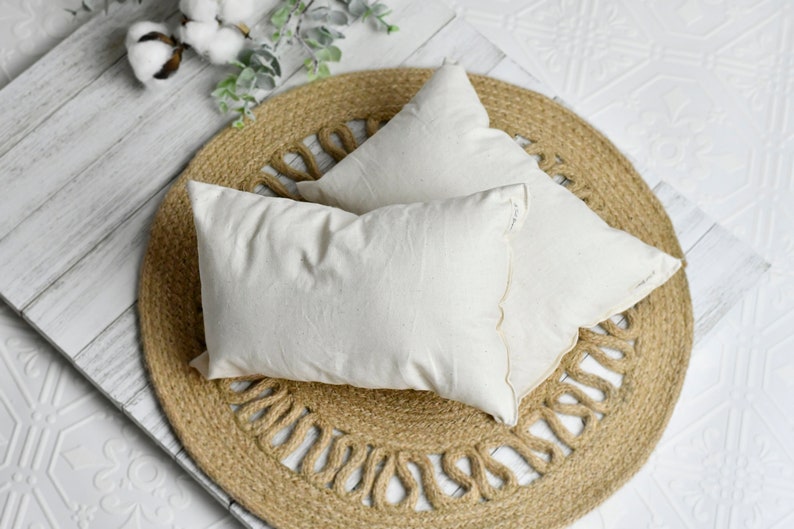 May include: Two off-white rectangular pillows rest on a round jute rug with a decorative edge. The pillows are made of a textured fabric. Cotton sprigs and greenery are in the background. The pillows are on a white wooden surface.