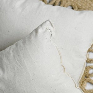 May include: Two off-white rectangular pillows with scalloped edges are displayed. The pillows are made of a textured fabric and rest on a woven jute rug. The corner of one pillow has a small printed text.