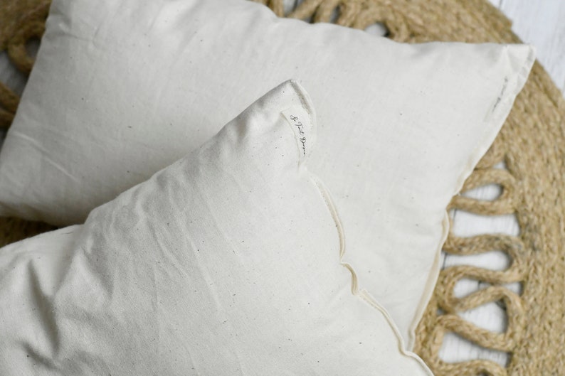May include: Two off-white rectangular pillows with scalloped edges are displayed. The pillows are made of a textured fabric and rest on a woven jute rug. The corner of one pillow has a small printed text.
