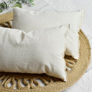 May include: Two rectangular, off-white pillows with a textured, linen-like appearance. They are placed on a round, woven jute mat. The pillows have a simple, minimalist design, suggesting a focus on natural materials and neutral tones.