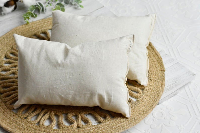 May include: Two rectangular, off-white pillows with a textured, linen-like appearance. They are placed on a round, woven jute mat. The pillows have a simple, minimalist design, suggesting a focus on natural materials and neutral tones.