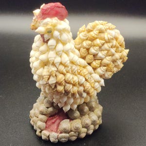 May include: A decorative rooster figurine crafted from various small seashells. The body is composed of white and tan spiral shells, featuring a prominent red shell crest on the head and a base covered in grey, rounded, textured seashells.