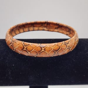 May include: A circular bangle bracelet featuring a textured, repeating diamond-shaped pattern in shades of tan, brown, and dark brown. The surface of the bracelet has a glossy finish and displays a distinct scale-like appearance throughout the exterior.