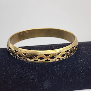 May include: A gold-toned metal bangle bracelet featuring a continuous openwork lattice pattern of intersecting diamond shapes. The bracelet has a smooth, polished finish along the top and bottom edges, framing the intricate geometric cutout design.
