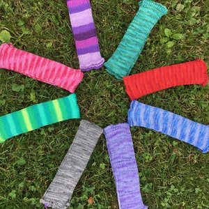 May include: A collection of eight knitted arm warmers in various colors, including pink, purple, green, red, and blue. Each arm warmer has a ruffled cuff and is arranged in a circular pattern on a bed of green grass.