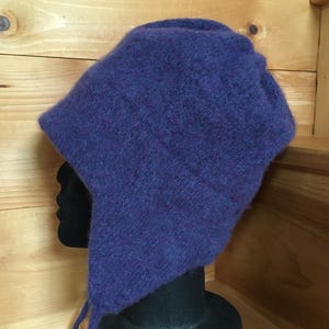 May include: A deep purple, fuzzy knit hat with ear flaps and dangling ties. The hat features a textured, soft-looking surface with subtle variations in the purple yarn color, providing full coverage for the head and ears in a classic winter style.