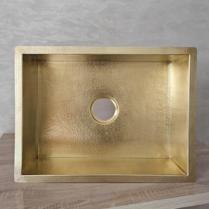 Puede incluir: Un fregadero rectangular con un acabado dorado metálico y una textura martillada distintiva en toda su superficie interior. El lavabo cuenta con una abertura de drenaje circular centrada y paredes verticales rectas con un borde ancho y plano.