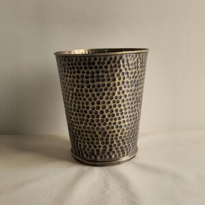 May include: A cylindrical metal vessel featuring a dark, hammered exterior finish with a repeating dimpled texture. The rim and base are defined by smooth, polished metallic bands that contrast with the textured body of the container.