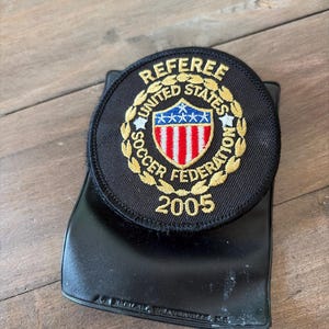 May include: A circular black embroidered patch featuring the text REFEREE UNITED STATES SOCCER FEDERATION and the year 2005 in gold thread. The center displays a shield with red and white vertical stripes and blue stars, surrounded by a gold wreath.