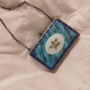 May include: A rectangular metal clutch with a turquoise marbled enamel finish and a dark blue border. The center features an oval white guilloche panel with a small floral motif of pink, yellow, and blue flowers. It includes a thin metal chain.