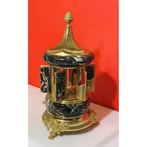 May include: A decorative carousel-style music box featuring black and white marble panels, a polished gold-tone metal frame, and a pointed finial top. The structure includes hinged panels and ornate gold feet, showcasing a central rotating figure.