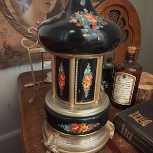 May include: A black carousel-style music box with a glossy finish, featuring gold-tone accents and colorful floral decals on the dome and base. The cylindrical body is supported by gold pillars, and the base rests on three ornate, leaf-shaped feet.
