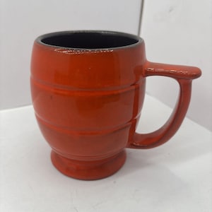 May include: A glossy, barrel-shaped ceramic mug with a vibrant orange exterior glaze and a dark, contrasting interior. The mug features three horizontal embossed bands around the body, a sturdy side handle, and a raised pedestal base.