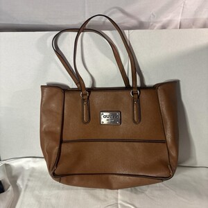 May include: A brown tote bag with two shoulder straps and a rectangular silver-toned metal plaque on the front center. The plaque features the text GUESS in embossed lettering. The bag has a structured base and a smooth, solid-colored exterior.