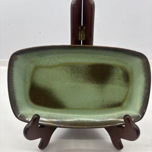 May include: A rectangular ceramic tray featuring a gradient glaze that transitions from a light sage green center to dark brown edges. The surface has a mottled, textured appearance with darker brown patches blended into the green finish.