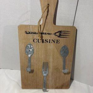 May include: A light brown wooden wall hook board shaped like a cutting board with a jute hanging string. It features a black printed fork graphic, the word CUISINE, and three metal hooks shaped like two spoons and one fork attached to the front.