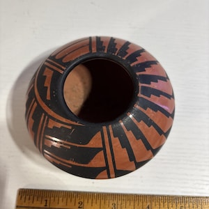 May include: A round terracotta-colored pottery bowl featuring black geometric patterns and a wide circular opening. A wooden ruler placed at the base shows the object measures approximately 3.5 inches in width at its widest point.