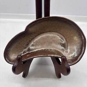 May include: A kidney-shaped ceramic dish featuring a glossy glaze with a gradient of dark brown along the outer edges transitioning to a lighter, speckled beige tone in the center. The dish has a smooth, curved rim and a shallow, concave basin.