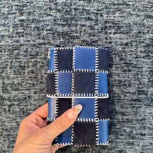 Pode incluir: Uma capa de caderno retangular com um padrão xadrez composto por quadrados de couro azul liso e quadrados de pele com pelo azul-marinho escuro. Os quadrados individuais são unidos por costuras brancas grossas e visíveis nas bordas.