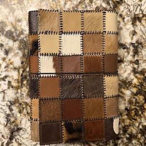 Könnte beinhalten: Ein rechteckiger Notizbuchumschlag aus einem Patchwork aus quadratischen Leder- und Fellstücken in verschiedenen Braun-, Beige-, Creme- und Schwarztönen. Die Quadrate sind mit dunkelbraunen Überwendlichstichen verbunden und durch ein dünnes schwarzes Gummiband gesichert.