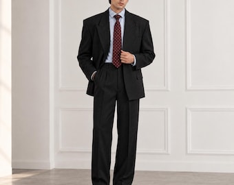 Può includere: Un abito grigio antracite scuro composto da un blazer strutturato e pantaloni abbinati con pinces. L'insieme è abbinato a una camicia a righe azzurre e bianche e a una cravatta rosso scuro con un piccolo motivo ripetuto.