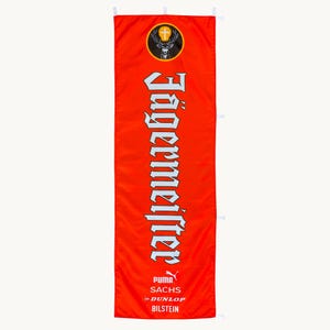 Puede incluir: Una pancarta rectangular vertical de color naranja con el logotipo de Jägermeister en la parte superior, que incluye una cabeza de ciervo y una cruz. La palabra Jägermeister está impresa verticalmente en letras góticas blancas, seguida de los logotipos de Puma, Sachs, Dunlop y Bilstein.