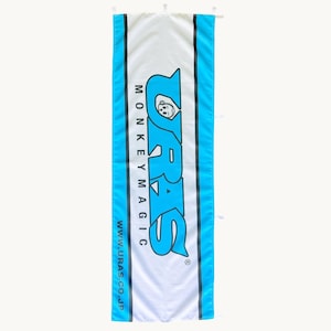 Op de afbeelding: Een rechthoekige verticale banner met een wit middenpaneel geflankeerd door felblauwe zijranden. De tekst URAS MONKEY MAGIC is gedrukt in grote blauwe letters met een kleine aapafbeelding, en de URL WWW.URAS.CO.JP is in het zwart gedrukt.