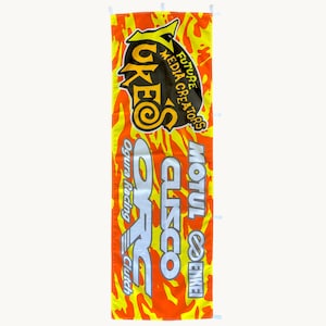 Op de afbeelding: Een verticaal rechthoekig spandoek met een levendige oranje en gele vlammenpatroon achtergrond. Het spandoek toont de tekst YUKES FUTURE MEDIA CREATORS, ORC OGURA RACING CLUTCH, CUSCO, MOTUL en ENKEI in dikke zwarte, witte en zilveren letters.