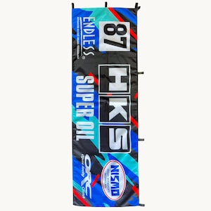 Peut inclure: Une bannière rectangulaire avec un motif abstrait noir, bleu et sarcelle. Le texte indique ENDLESS, 87, HKS, SUPER OIL, ORC Ogura Racing Clutch et NISMO Nissan Motorsports International. La bannière possède des languettes en tissu noir.