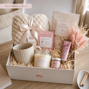 Peut inclure: Un coffret cadeau contenant une couverture en tricot crème nouée d'un ruban rose, une tasse en céramique côtelée, une bougie en pot, un sachet de sels de bain, un sachet de thé fraise-vanille, une crème pour les mains et des fleurs séchées roses.