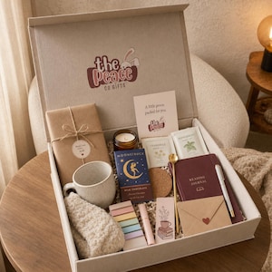 Könnte beinhalten: Eine Geschenkbox von The Peace Co Gifts mit einem verpackten Buch, einer gesprenkelten Keramiktasse, beigen flauschigen Socken, einer Moonstruck-Milchschokoladentafel, Teepäckchen, einem weinroten Lesetagebuch, einem goldenen Löffel, einem Stift und einem kleinen Umschlag.