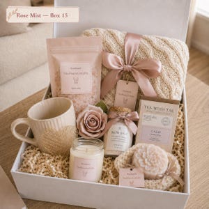 May include: A gift box containing a cream-coloured cable knit blanket tied with a dusty rose ribbon, a beige ribbed mug with a gold heart, a pouch of bath salts, a glass jar of bath salts, a rose, a box of Tea With Tae, and a floral-shaped soap.