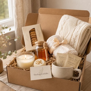 Könnte beinhalten: Eine Geschenkschachtel aus Pappe mit einer cremefarbenen Zopfstrickdecke, einem Glas Honig mit Holzlöffel, einer Keksschachtel, einer gerippten Glaskerze, einer kleinen runden Seife, zwei Kräuterteebeuteln und einer Karte mit der Aufschrift ThePeaceCoGifts.