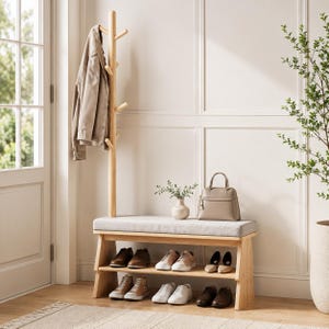 May include: A light wood hall tree featuring a vertical coat rack with multiple pegs, a bench with a light gray upholstered seat, and two lower shelves holding various pairs of shoes. A beige jacket hangs on the rack and a handbag sits on the bench.