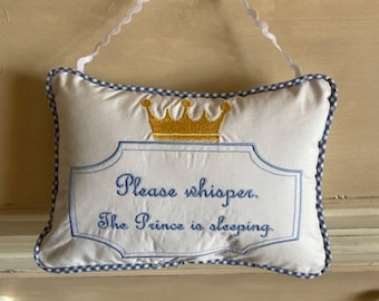 Könnte beinhalten: Ein rechteckiges weißes Stoffschild mit blau-weißem Vichy-Karo-Rand und einem weißen, gewellten Band zum Aufhängen. Das Schild zeigt eine goldene Stickkrone über dem blauen Text: Please whisper. The Prince is sleeping. Das Schild ist zentriert.