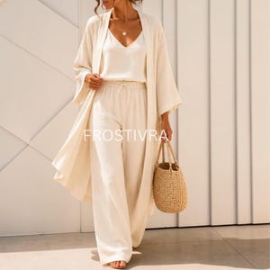 May include: A cream-colored three-piece outfit consisting of a sleeveless V-neck top, wide-leg trousers with a drawstring waist, and an open-front long-sleeve duster cardigan. The set is paired with a woven straw tote bag. The text FROSTIVRA is visible.