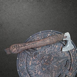 May include: A dark brown wooden axe with intricate carvings on the handle and a silver metal head featuring engraved patterns, resting on a circular wooden shield decorated with a detailed relief of a wolf and knotwork designs.