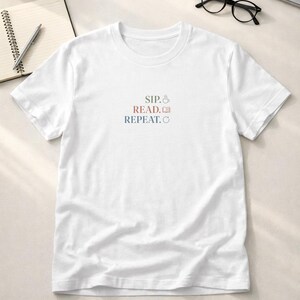 May include: A white short-sleeved t-shirt featuring the text SIP. with a coffee cup icon, READ. with an open book icon, and REPEAT. with a circular arrow icon printed in the center of the chest in muted brown, red, and blue colors.