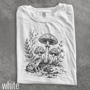 May include: A white short-sleeve t-shirt featuring a black ink-style illustration of a frog sitting next to three mushrooms and leafy plants. The inner neck label displays the text Comfort Colors. The design is centered on the front of the shirt.