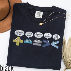 Op de afbeelding: Een zwart t-shirt met een afbeelding van vijf kleurrijke wiskundige symbolen met cartoonogen en tekstballonnen. De tekst luidt: Let's add it up!, Take it away., Sounds fair., I'm bigger., en Not quite. Het t-shirt heeft een Comfort Colors label.