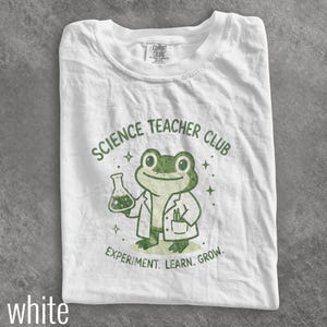 May include: A white t-shirt featuring a green graphic of a frog wearing a lab coat, holding a beaker, and surrounded by small star icons. The text reads SCIENCE TEACHER CLUB and EXPERIMENT. LEARN. GROW. in a dark green, distressed font style.