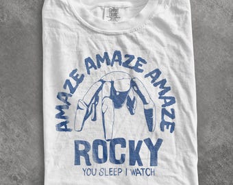 May include: A white short-sleeve t-shirt featuring a blue graphic of a multi-legged creature. The text reads "AMAZE AMAZE AMAZE" in an arc above, "ROCKY" in large letters below, and "YOU SLEEP I WATCH" at the bottom. The shirt has a crew neckline.