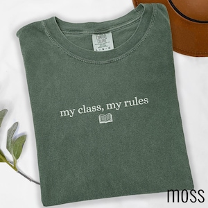 May include: A moss green crewneck t-shirt featuring the embroidered white text "my class, my rules" above a small open book icon. The shirt has a Comfort Colors brand label at the inner neckline indicating a size large and 100% cotton material.