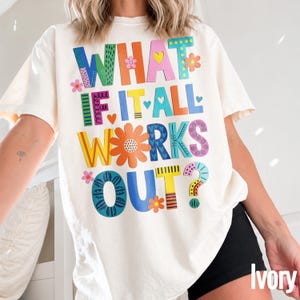 Może przedstawiać: T-shirt z krótkim rękawem w kolorze kości słoniowej z kolorowym napisem WHAT IF IT ALL WORKS OUT? w różnych czcionkach i wzorach. Projekt zawiera elementy dekoracyjne, takie jak małe kwiatki, serca i kropki w odcieniach niebieskiego, pomarańczowego, różowego i żółtego.