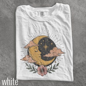 May include: A white t-shirt featuring a graphic print of a yellow crescent moon with a face, surrounded by pink clouds, a dark blue starry sky, and a single pink flower with green leaves at the bottom. The inner neck label reads Comfort Colors.