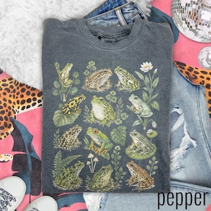 May include: A charcoal gray crewneck t-shirt featuring a detailed graphic print of various frogs and toads in shades of green, brown, and yellow, interspersed with botanical illustrations of leaves, ferns, and small white flowers.
