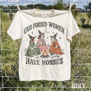 Puede incluir: Una camiseta de manga corta color marfil con un estampado gráfico de tres figuras con sombreros de bruja sentadas alrededor de una fogata. El texto dice GOD FORBID WOMEN HAVE HOBBIES. La ilustración incluye una luna creciente, estrellas y ropa de color verde, rosa y naranja.