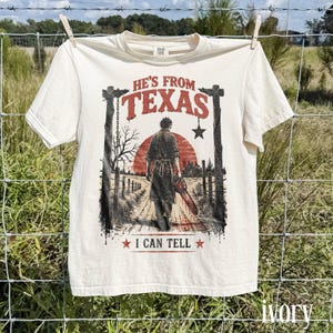 Pode incluir: Uma t-shirt de manga curta na cor marfim com uma estampa gráfica de uma pessoa segurando uma motosserra em uma paisagem rural. O texto diz HE'S FROM TEXAS e I CAN TELL, acompanhado por um sol vermelho, postes de madeira e uma estrela.