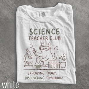 Peut inclure: Un t-shirt blanc à manches courtes avec un motif de chat portant des lunettes de protection. Le texte indique SCIENCE TEACHER CLUB et EXPLORING TODAY, DISCOVERING TOMORROW. Le dessin inclut un microscope, un flacon, des éprouvettes et une plante.