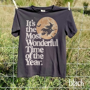 May include: A charcoal gray t-shirt featuring a graphic of a witch flying on a broomstick in front of a large, textured orange moon. The text "It's the Most Wonderful Time of the Year" is printed in a distressed white font alongside small bat silhouettes.